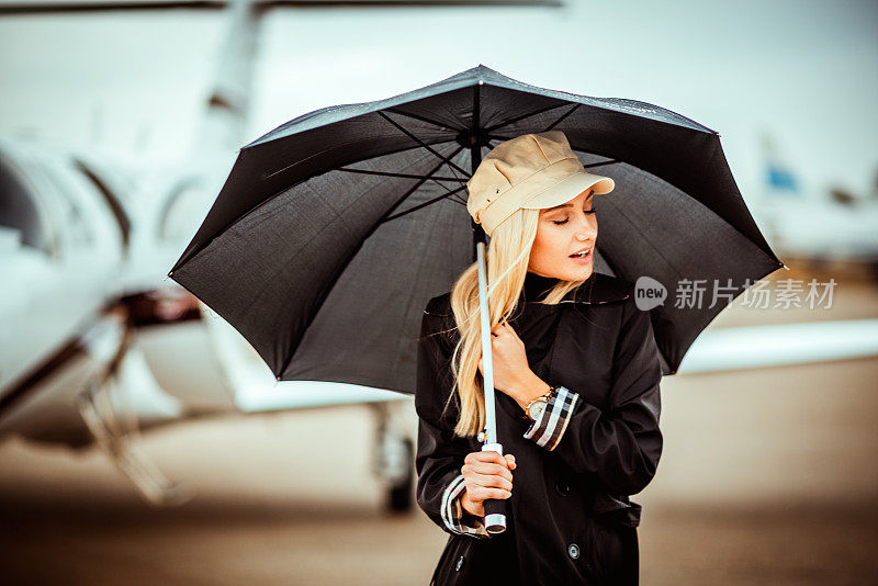 一位年轻漂亮的金发女郎撑着一把黑伞站在一架停在机场跑道上的私人飞机前