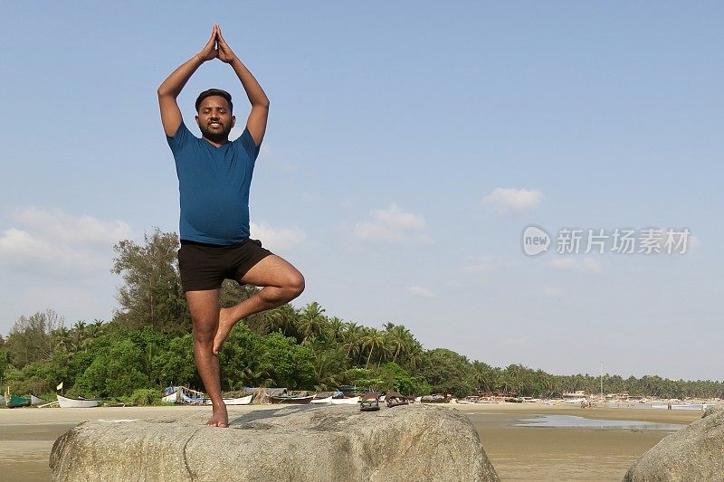 印度人在岩石上练习树姿势(Vrksasana)瑜伽姿势，水的边缘，帕洛伦海滩，果阿，印度