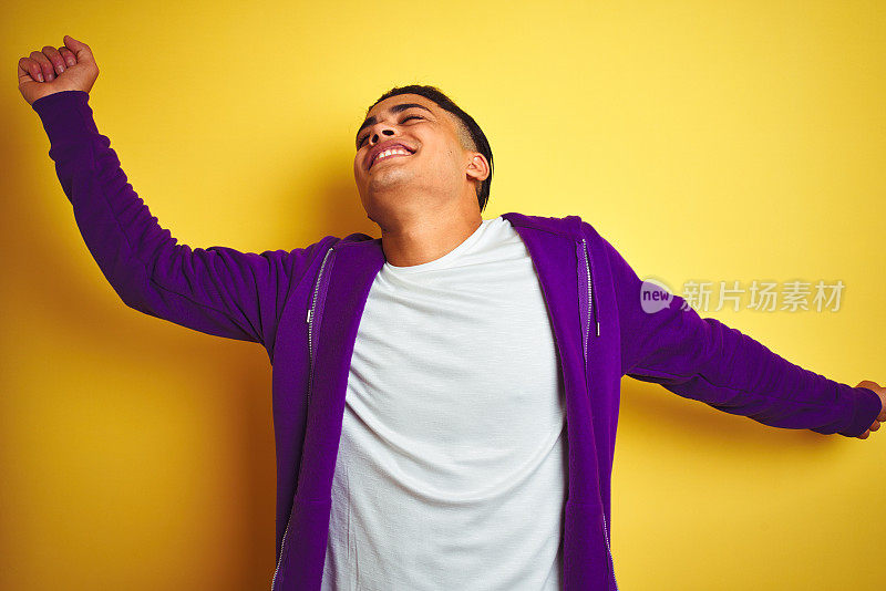 年轻的巴西男子穿着紫色的运动衫站在孤立的黄色背景跳舞快乐和欢快，微笑着移动随意和自信听音乐