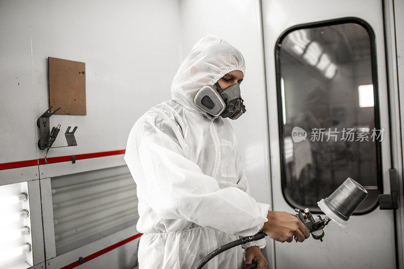 专业汽车油漆工一边穿着工作服，一边拿着呼吸面罩拿着喷枪在喷漆间里