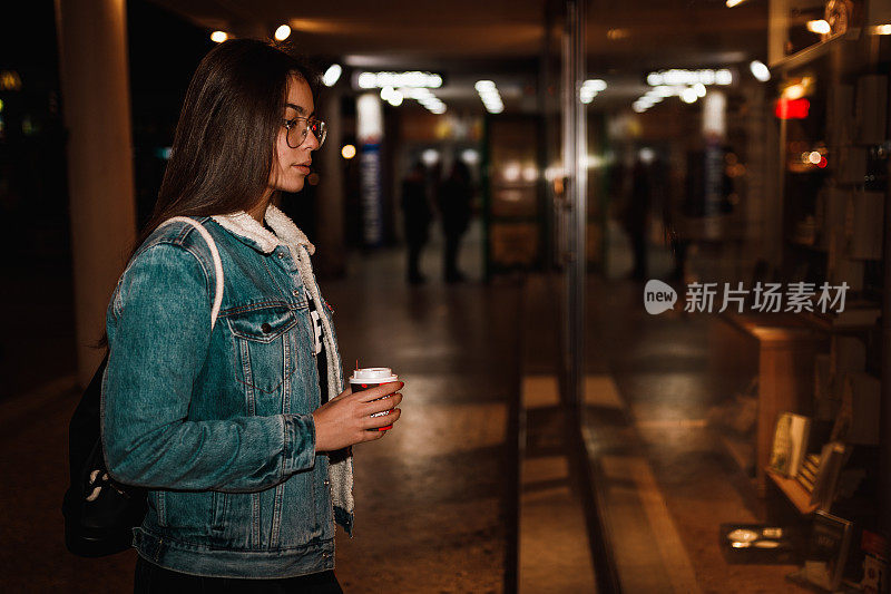 一个十几岁的女孩晚上拿着咖啡杯站在商店的橱窗前