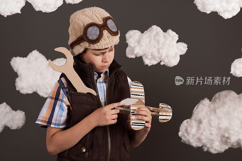 一个男孩在玩纸板飞机和扳手，梦想成为一名飞行员，穿着复古风格的夹克，戴着头盔和眼镜，棉絮云，灰色的背景，色调为棕色