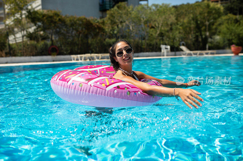 在一个炎热的夏天，一个美丽的女孩在充气甜甜圈上的游泳池里玩得很开心