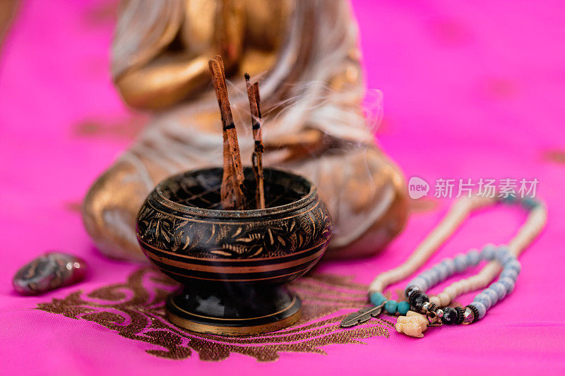 在一个没有聚焦的金佛前面的香碗里，靠近熏香的香烛，还有一条美丽的粉红色印度教纱丽绳，上面有刺绣和印度珠宝