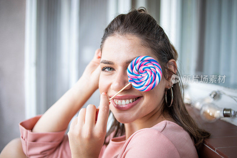 一位身穿粉红色连衣裙的美丽年轻女子坐在阳台上，手里拿着一根大棒棒糖。