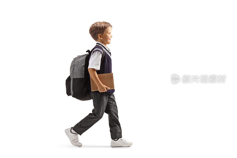 一个穿着制服的小男孩背着书包和一本书，走着