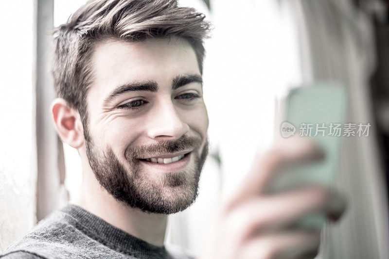 这是一幅带着胡子微笑的年轻人拿着智能手机的偷拍照片