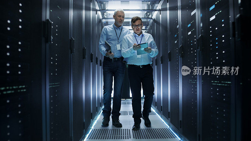 在数据中心，两个IT工程师穿过一排排的服务器机架。他们在平板电脑和笔记本电脑上工作。