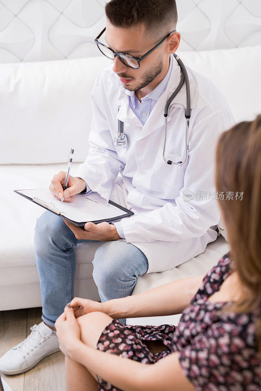 严肃忙碌的年轻男医生，戴着眼镜，穿着白大褂，坐在沙发上，一边保存医疗记录，一边与病人进行咨询