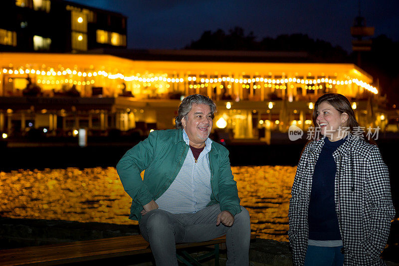 在蓝色的时间里，一对情侣在湖边摆姿势，以湖上的灯光为背景