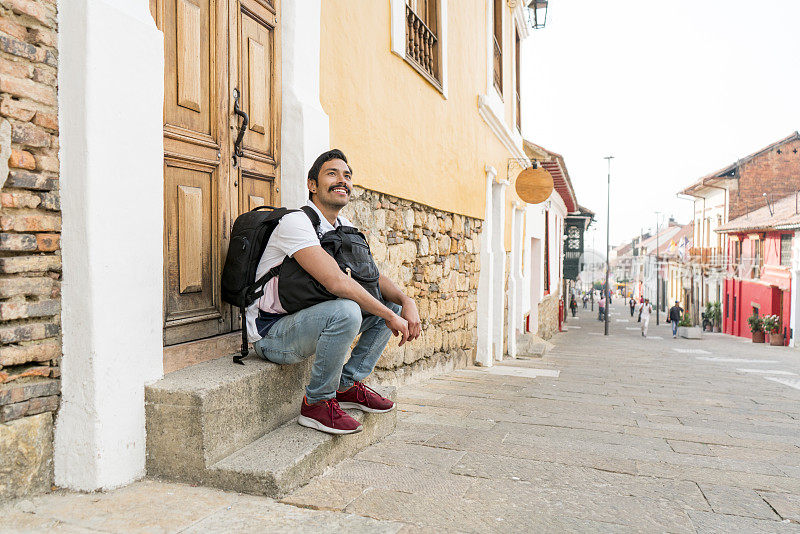 一名25岁的拉丁裔男子独自穿过波哥大的街道，他会在一所老房子门前坐上片刻
