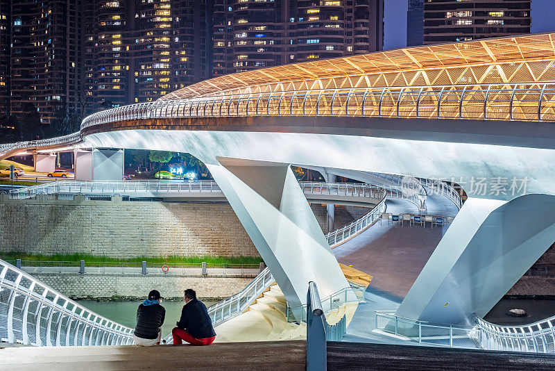 两个人坐在一座现代化的桥上，桥上的灯光在夜里照亮了河