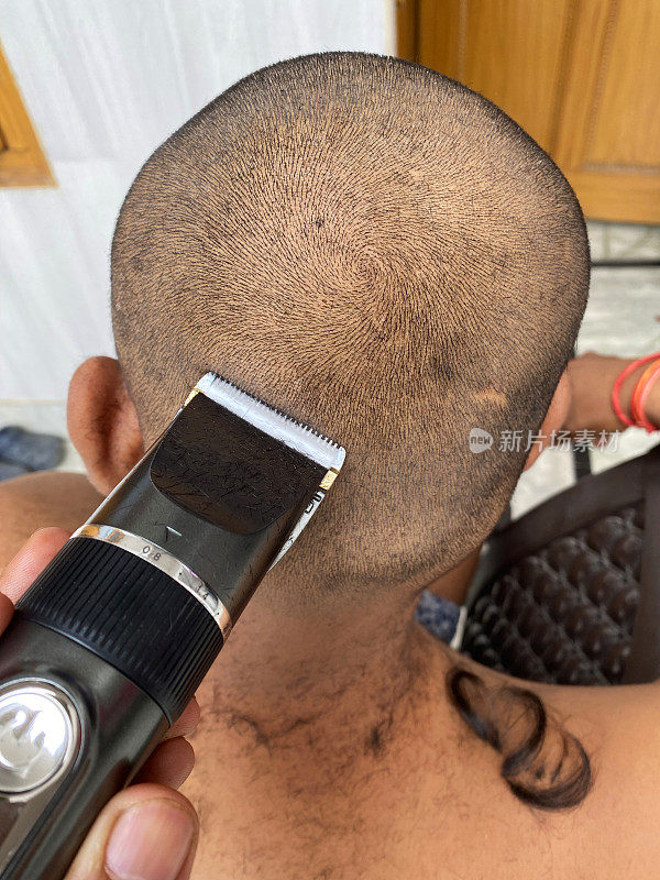 照片中赤裸上身的印度男子在家理发，电动剃须刀，理发师用剪发器剃短后背和侧面，后视镜，聚焦前景