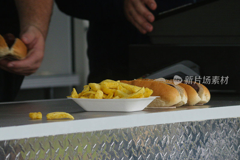 照片中，一群不知名的小贩在餐车柜台后面供应快餐外卖，用一次性聚苯乙烯盘子盛着薯条，用面包包着两个热狗香肠