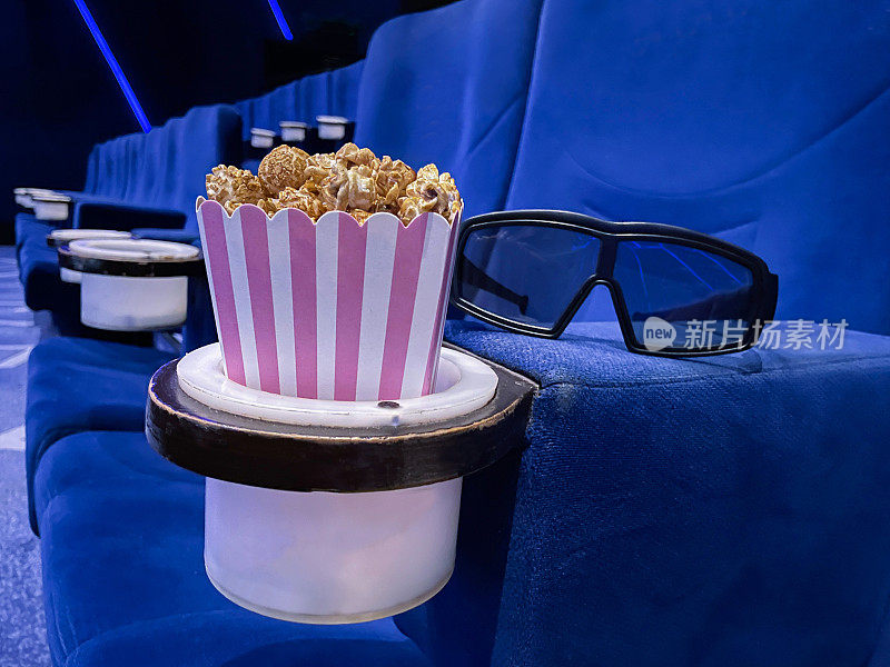 电影院里的爆米花盒图像，除了3D眼镜，电影零食盒，蓝色天鹅绒座椅，重点在前景