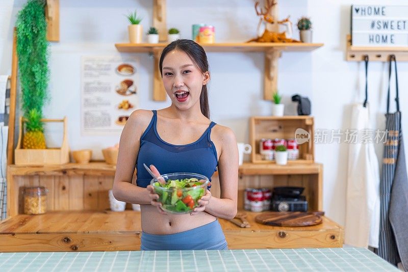 照片年轻美丽的亚洲女性女士在运动服高兴地炫耀一碗沙拉与樱桃番茄在她的房子的厨房节食和减肥健康的生活方式