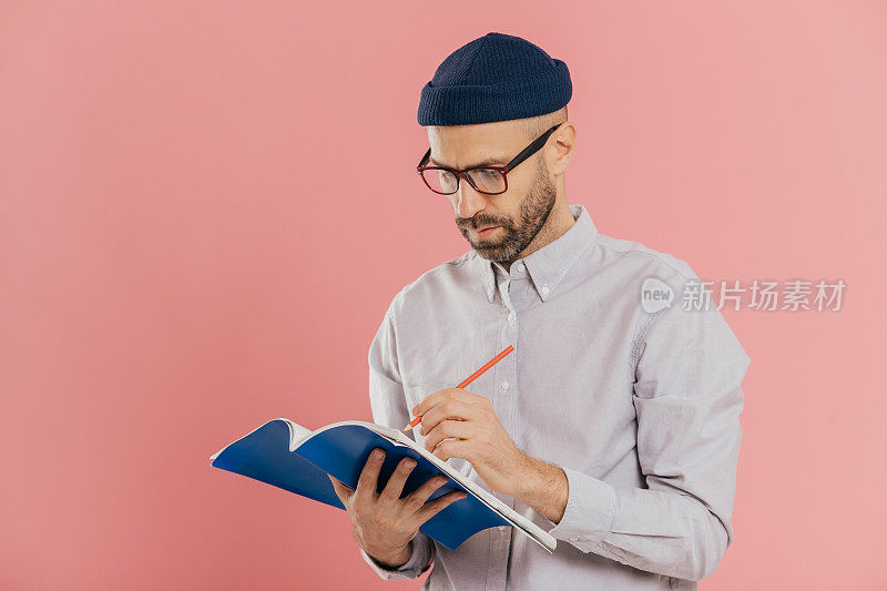 严肃的男性作家在记事本上写文章，有灵感，戴眼镜和正装，为新书准备创意，对着粉红色的工作室墙壁做模特。留着胡子的男人