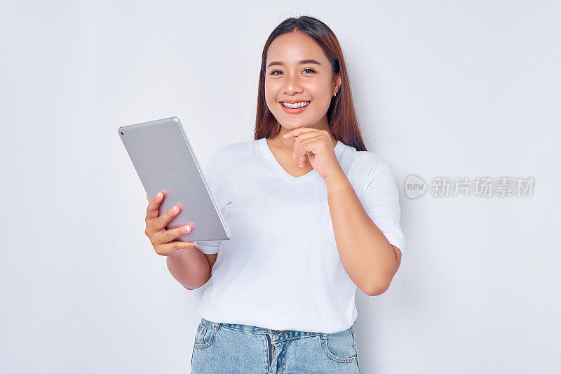 穿着白色t恤的年轻亚洲女孩用平板电脑微笑，用手触摸下巴，看着白色背景上孤立的相机