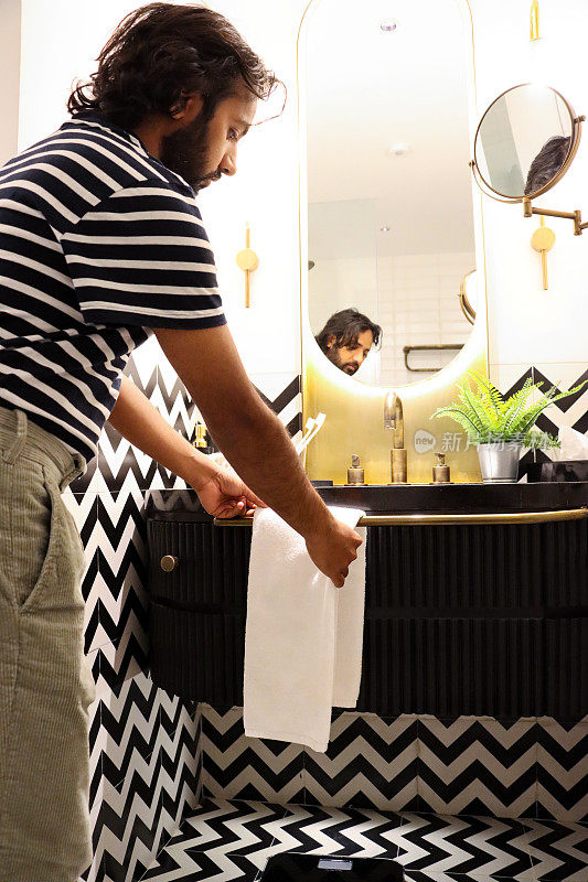 特写图像，印度男子站在浴室的水槽上，将折叠的白色毛巾挂在毛巾栏杆上，连接到洗手台单元，镜子里的倒影，人造蕨类植物在大理石浴室柜台上的金属锅里，专注于前景