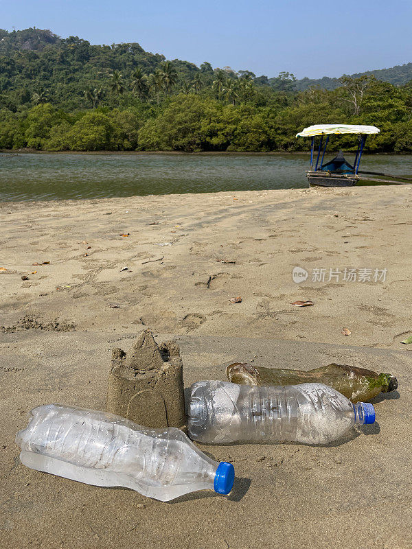 三个塑料水瓶周围的沙堡用桶和铲子在干燥，沙滩，海洋垃圾和污染的海岸，乱扔的沙子，渔船，林地棕榈树，重点在前景