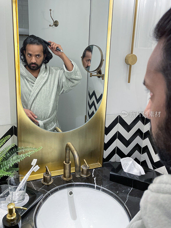 镜子里的印度男人穿着浴袍站在浴室水槽前，用梳子梳理凌乱潮湿的头发，自我护理和修饰的概念