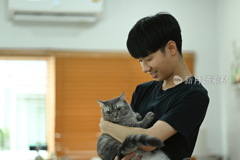 快乐的亚洲男人抱着他的猫站在客厅里开着空调。