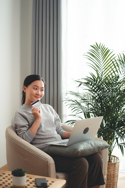 亚洲女人开心地微笑着坐在扶手椅上，用笔记本电脑和信用卡在家网上购物。