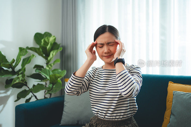 一名亚洲妇女正坐在家里客厅的沙发上饱受头痛之苦。