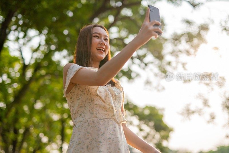 年轻的亚洲女子在公园用手机自拍。在郁郁葱葱的绿色森林里，美女视频通话