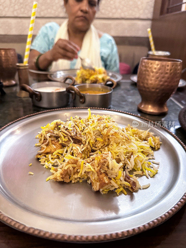 餐桌上的特写图像是金属盘子上的鸡肉印度香饭，藏红花皮饭，印度妇女穿着蓝色的萨尔瓦·卡米兹传统服装，白色的杜帕塔(围巾)背景，重点是前景