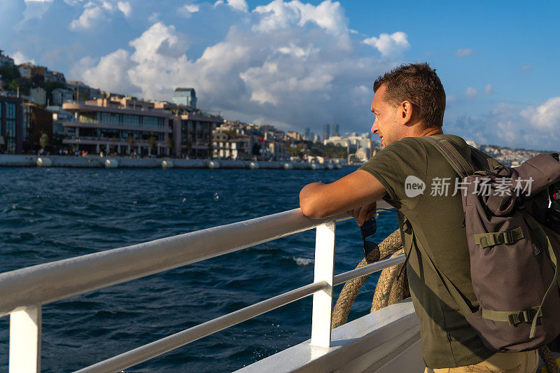 在一个阳光明媚的日子里，快乐的年轻男游客背着背包在伊斯坦布尔的渡轮上享受着大海和这座城市。旅游的概念，旅游