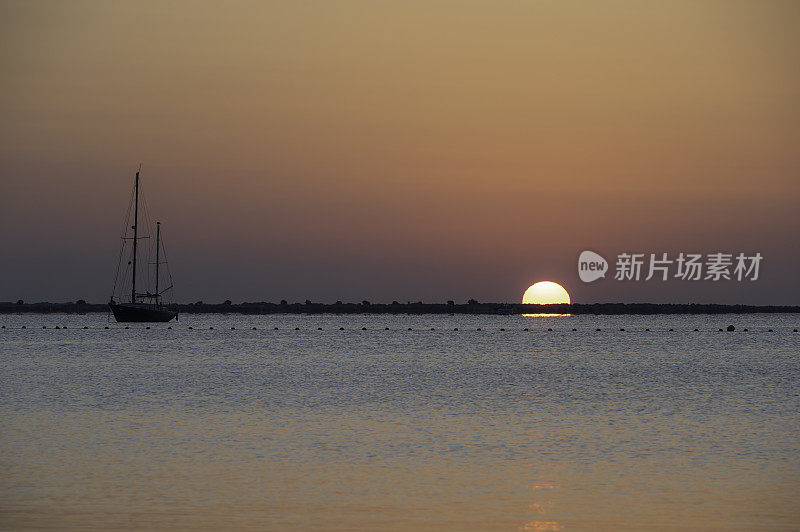 一轮黄色的太阳升上橘黄色的天空，在平静的地中海上空，在一艘单帆船旁边的水面上反射
