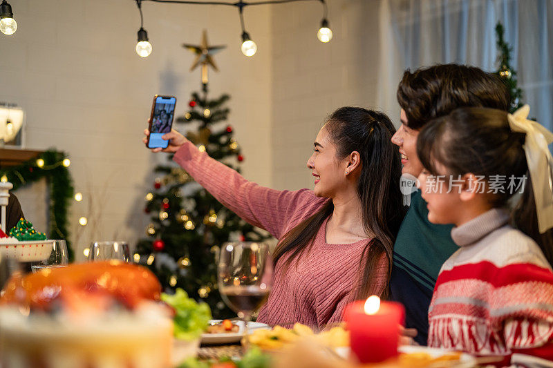 亚洲家庭在一起庆祝圣诞派对时拍照。年轻迷人的孩子女儿和父母在家里的餐桌上吃晚餐，享受节日感恩节、圣诞前夜的美食。