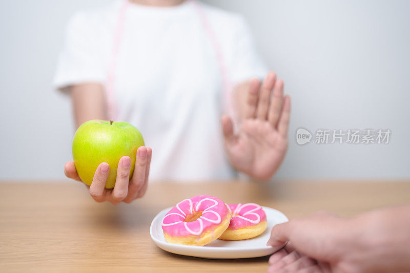女性手持青苹果而拒绝甜甜圈，女性健身在健康水果和不健康的垃圾食品之间选择。节食控制，体重减轻，肥胖，饮食方式和营养