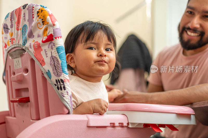 拉丁父亲给微笑的女儿喂奶，蹒跚学步的孩子在高脚椅上吃饭，爸爸和女儿在一个温暖的场景中亲密无间