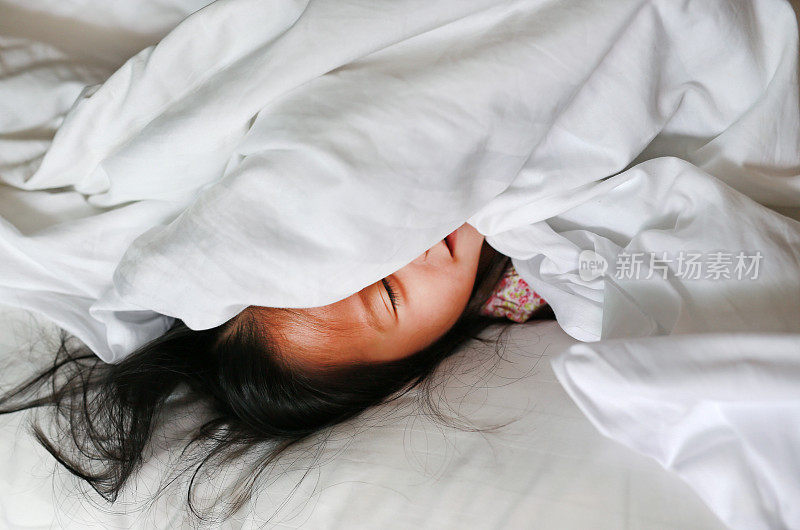 亚洲小女孩早上醒来，躺在床上的毯子下面。