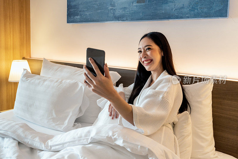 快乐的亚洲女人在床上用智能手机打视频电话。女游客在酒店里用智能手机。