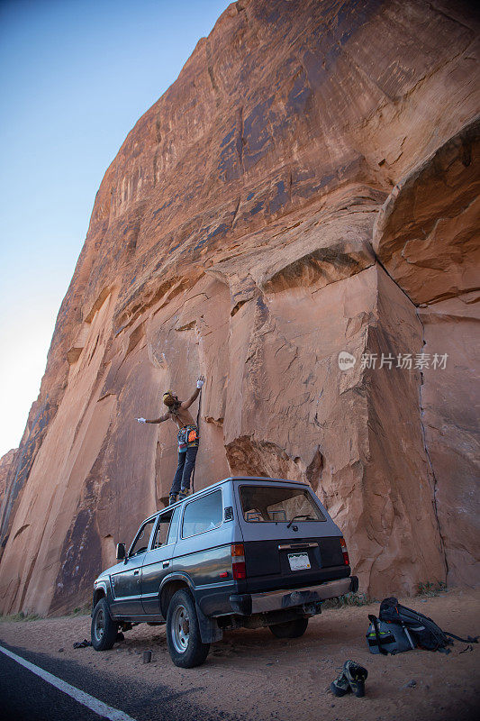 广角镜头。一名身穿攀岩装备的冒险男子站在一辆运动型多用途车顶上，双手举在空中，敬畏地看着巨大的峡谷岩面，准备在犹他州的摩押攀爬。
