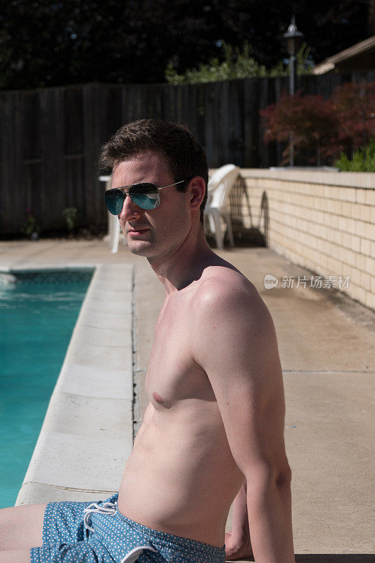 健康健康的男子坐在泳池边，脚在水里，没有衬衫和蓝色泳裤，戴着飞行员太阳镜。一个赤裸上身的运动员站在后院的泳池边。