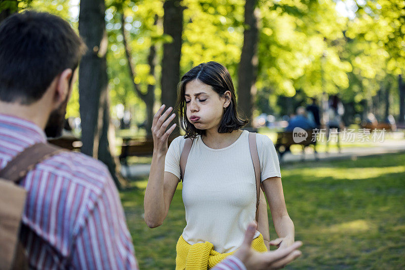 一对年轻夫妇在公园里吵架。一对情侣在一个有绿色背景的公园的户外严肃地交谈。夫妻争吵，对彼此不满意。