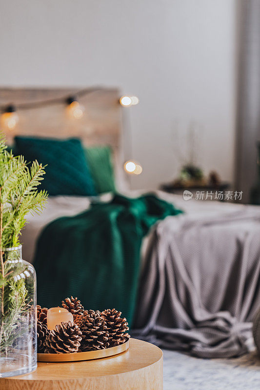 在优雅的卧室内部，咖啡桌上的玻璃花瓶和一盘球果和鲜花，灰色的床和翠绿色的床上用品