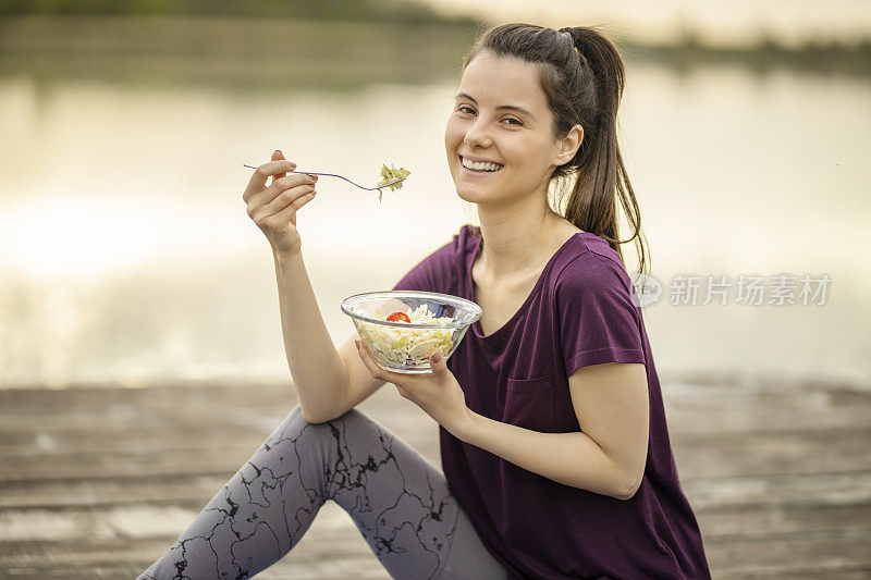 运动女性吃素食沙拉，坐在湖边码头上