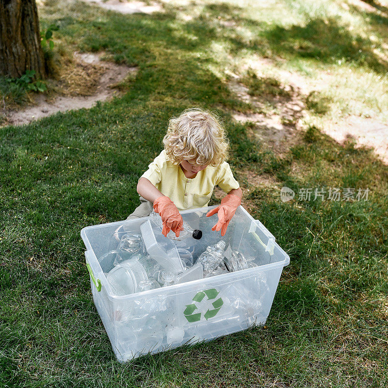 可爱的白人小男孩戴着橡胶手套，在森林或公园打扫时把塑料垃圾扔进回收站