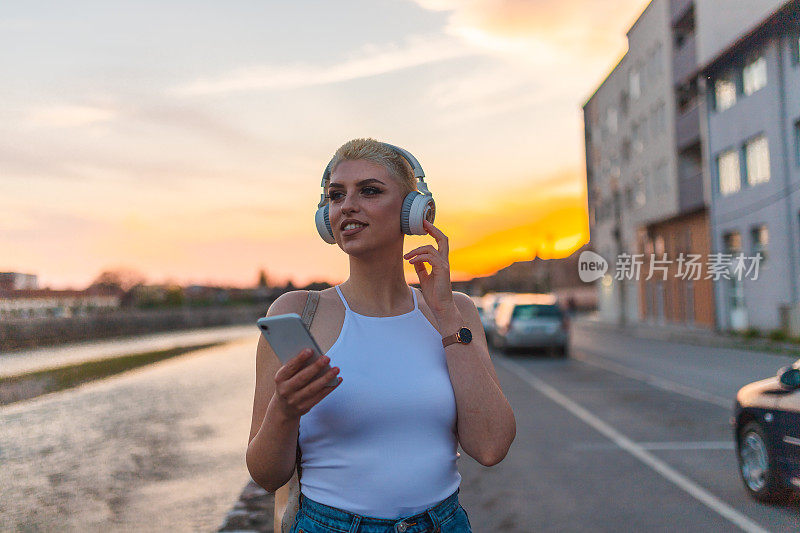 金发短发的年轻女子在夕阳下散步时戴着白色耳机听音乐