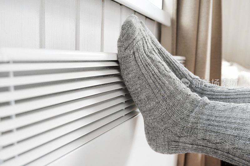 穿着针织袜的腿在电加热器上加热。冰冻的女人穿着温暖的羊毛袜子冻在冬天的寒冷。不舒服的呆在家里。女孩在现代暖气片上暖脚