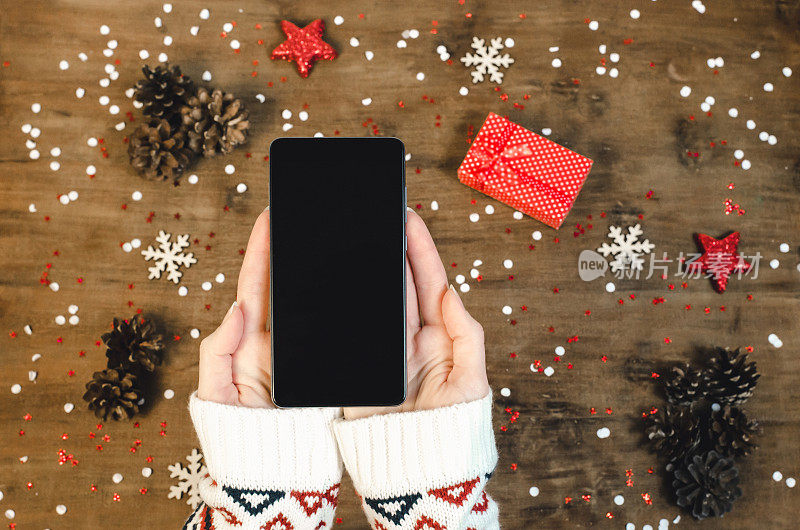 女性手握智能手机与oled显示屏上的木制背景与圣诞礼物雪花和雪花。新年快乐，帽子上的圣诞老人手机，平铺构图俯视图。
