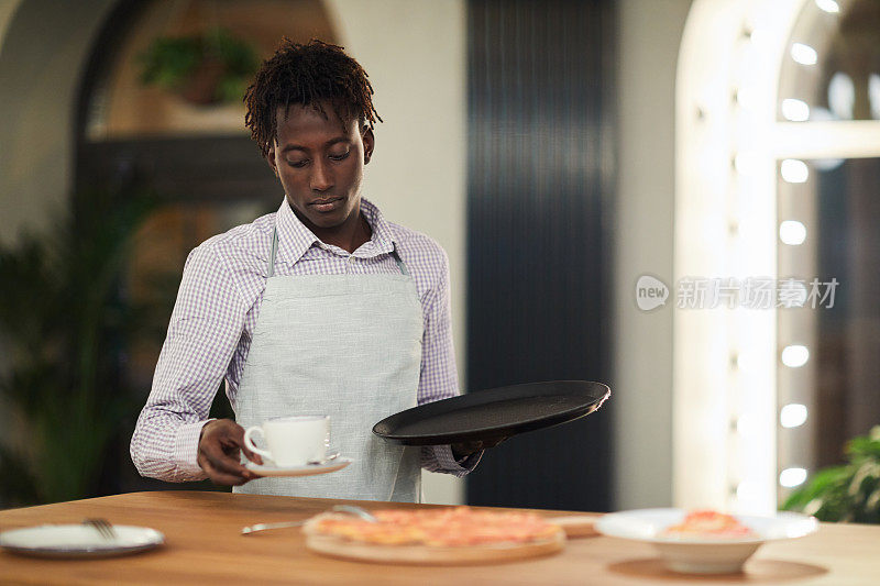 在现代餐厅里，年轻英俊的非洲裔美国人服务员系着围裙，端着盘子站在桌子旁，清理脏餐具