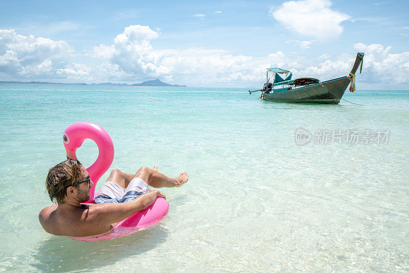 一个年轻人在田园般的海滩上放松，充气火烈鸟在清澈的海水中玩耍在泰国的岛屿。人们旅行奢侈，乐趣和酷的态度概念
