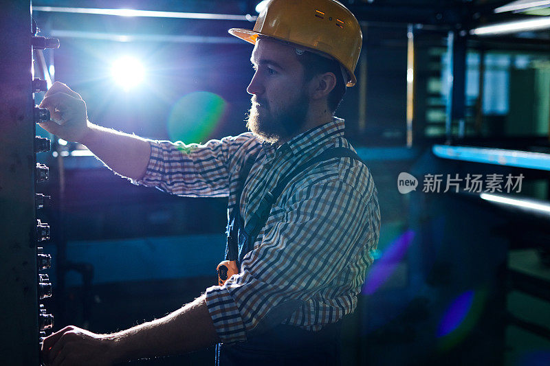 一名戴着安全帽、严肃忙碌的工程师，站在黑暗的车间的控制面板前，在工厂的机器上工作