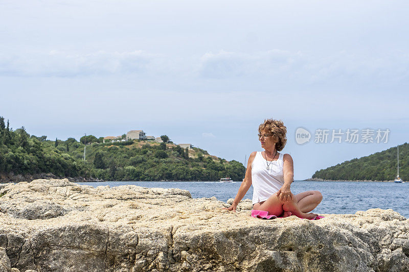 老女人坐在岩石海滩上练瑜伽